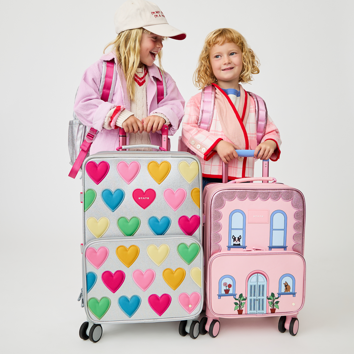 Two children standing behind a pink State Bags Mini Logan house-style rolling carry-on suitcase and a matching rolling suitcase covered in colorful hearts, wearing pink jackets with red trim and white caps.