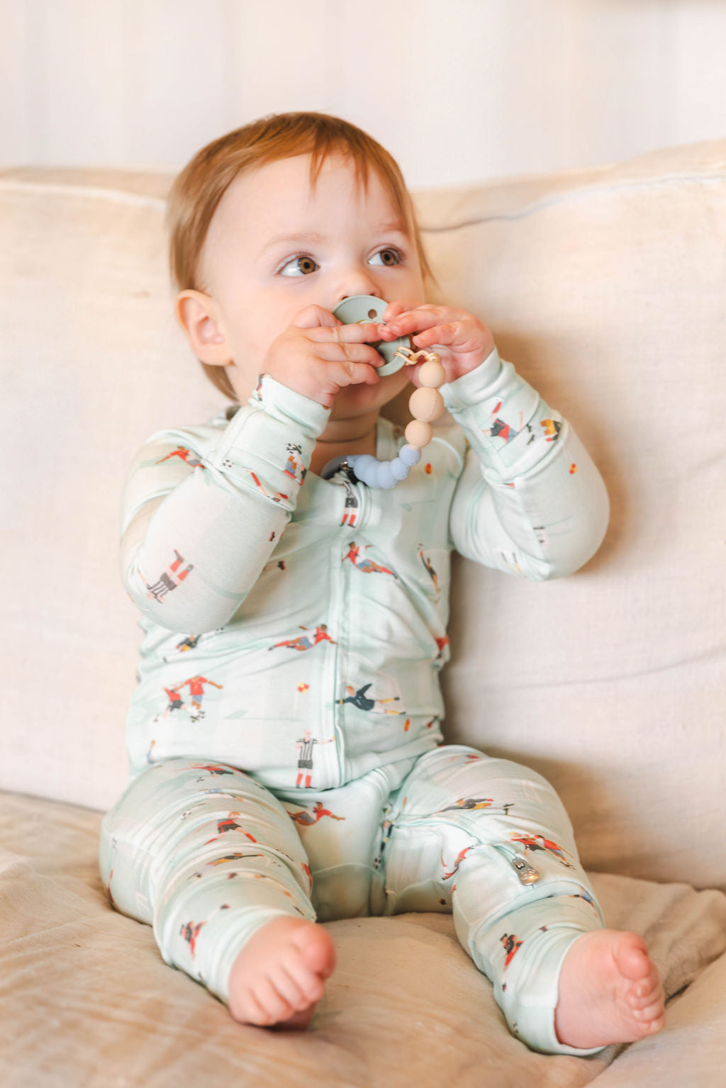 Baby in a light green onesie with bird pattern sitting on a beige couch.