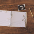 An open baby keepsake album with floral gold and beige borders lies on a wooden tabletop, alongside a small black-and-white ultrasound photo and a gold pen.