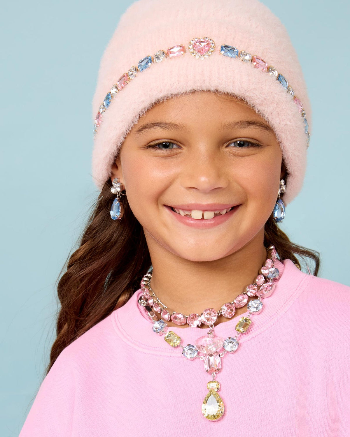 A young girl smiling while wearing a light pink fuzzy beanie with a jeweled headband, matching pink and blue teardrop earrings, and a pink and blue rhinestone necklace against a light blue background.