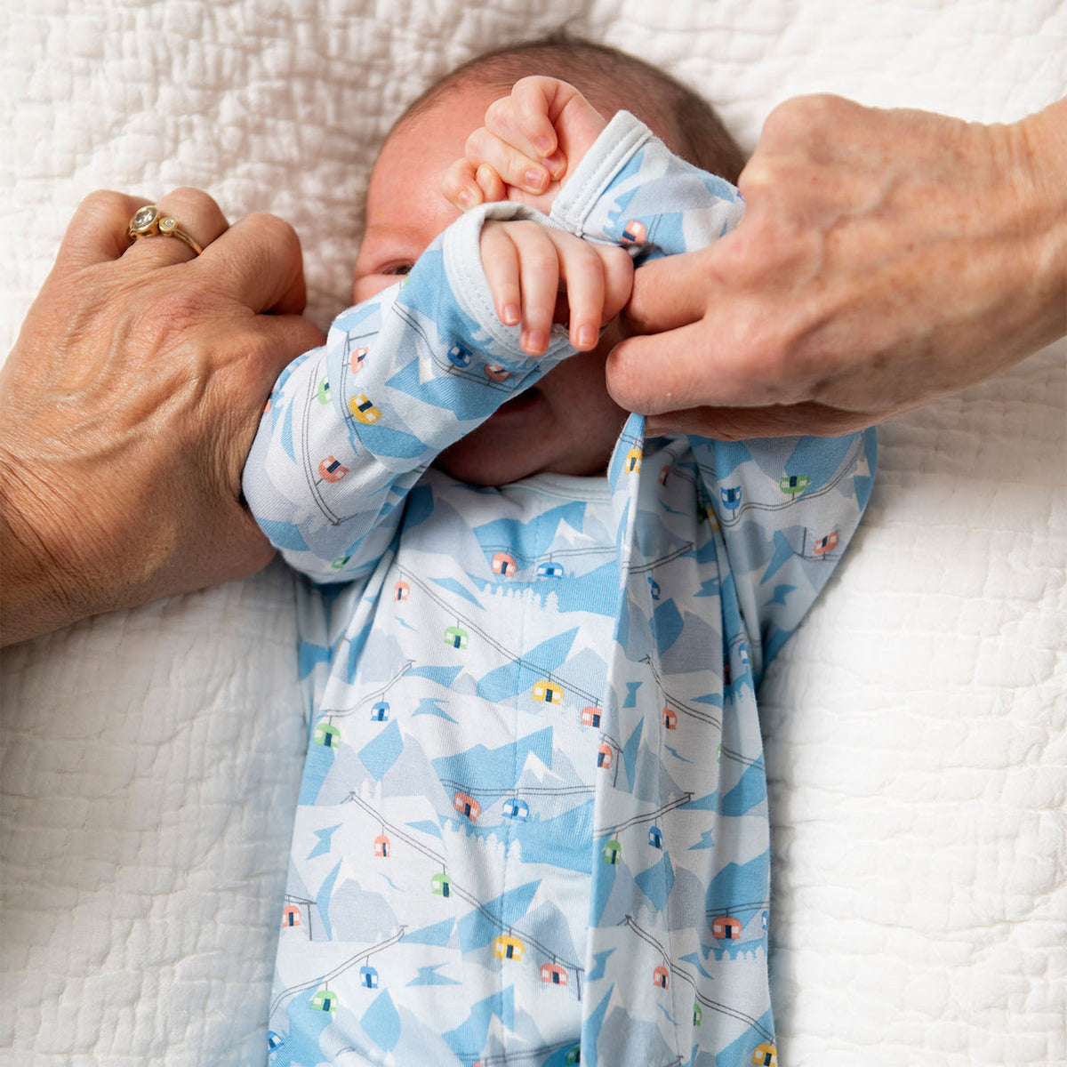 Baby in a blue airplane-patterned onesie being held by an adult on a white blanket.