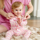 A baby wearing a pink long-sleeve footed pajama set with a small floral pattern sits on a fluffy rug while an adult in matching pink pajamas adjusts the baby's top.
