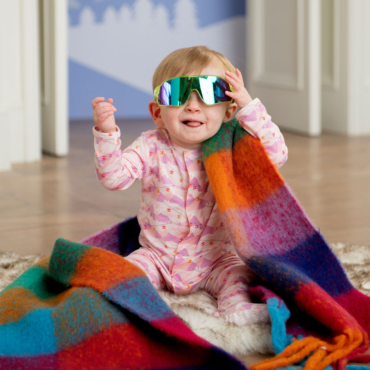 Child wearing colorful goggles and a pink outfit, sitting on a multicolored blanket indoors.