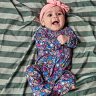 A baby lying on a green and white striped blanket, wearing a pink knotted headband and a long-sleeve footed sleeper with a colorful floral pattern.