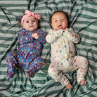 Two babies lying side by side on a green and white striped blanket, wearing matching floral and fruit-print footed onesies. The baby on the left wears a blue, purple, and pink floral one-piece with a pink bow headband.