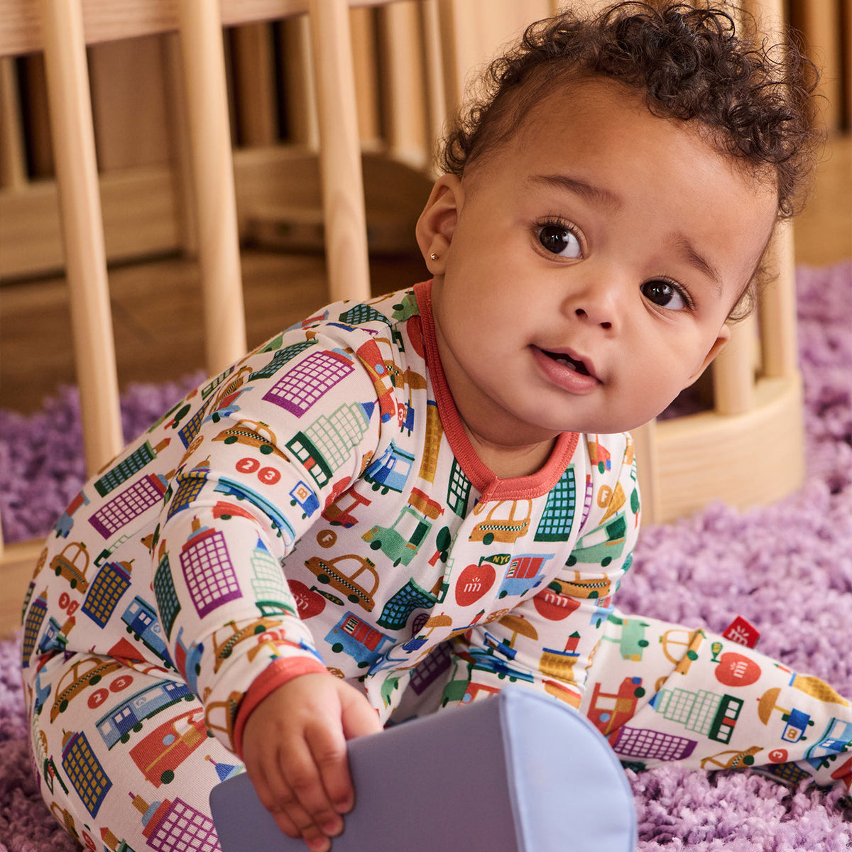 A baby wearing a colorful, long-sleeve onesie with a playful city-themed print of cars, buses, and buildings, sitting on a purple shag rug in front of a wooden crib.
