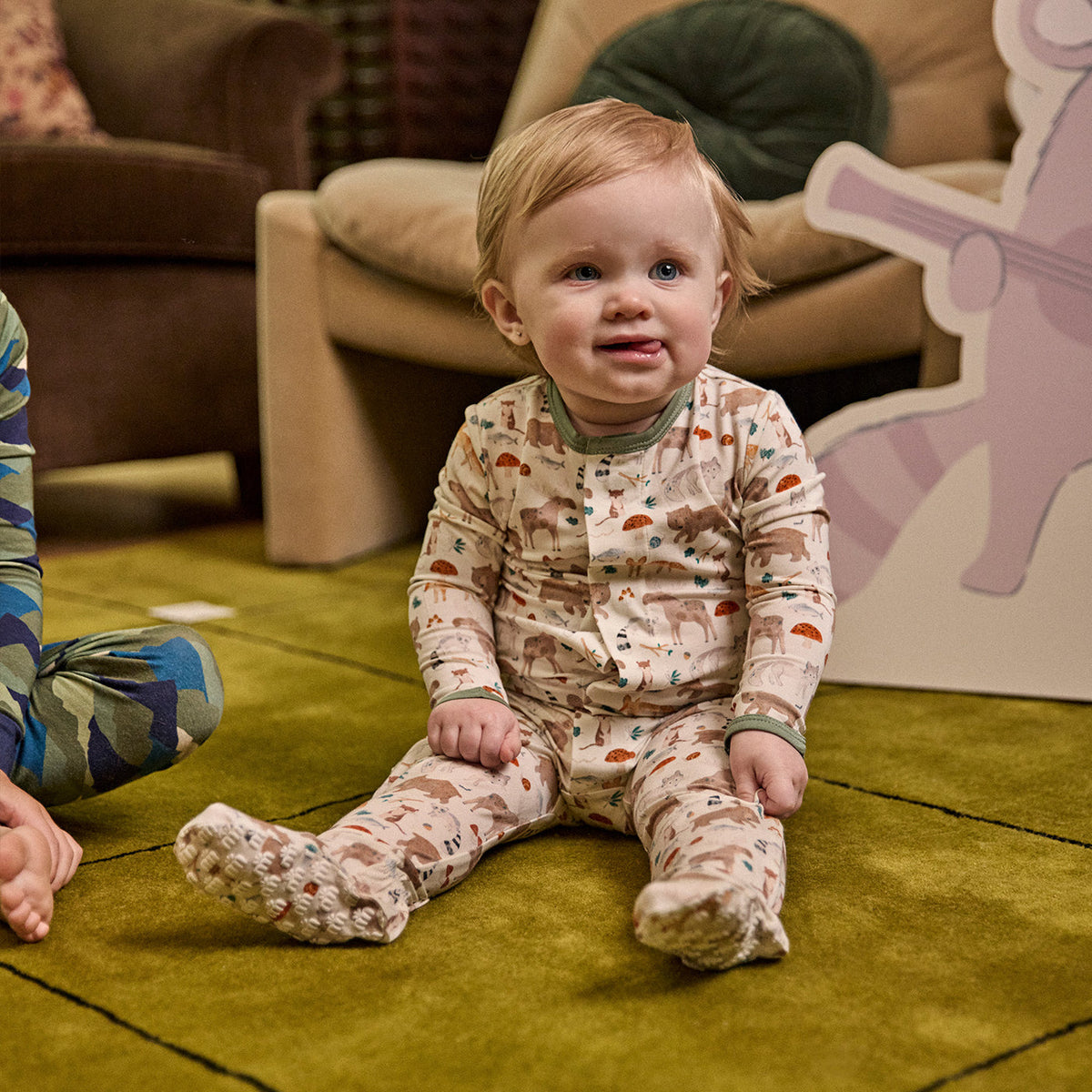 A baby sitting on a green carpet wearing a long-sleeve footed pajama romper with a woodland tree pattern. The background shows a cozy living room setting with a brown armchair and a large cartoon character cutout.
