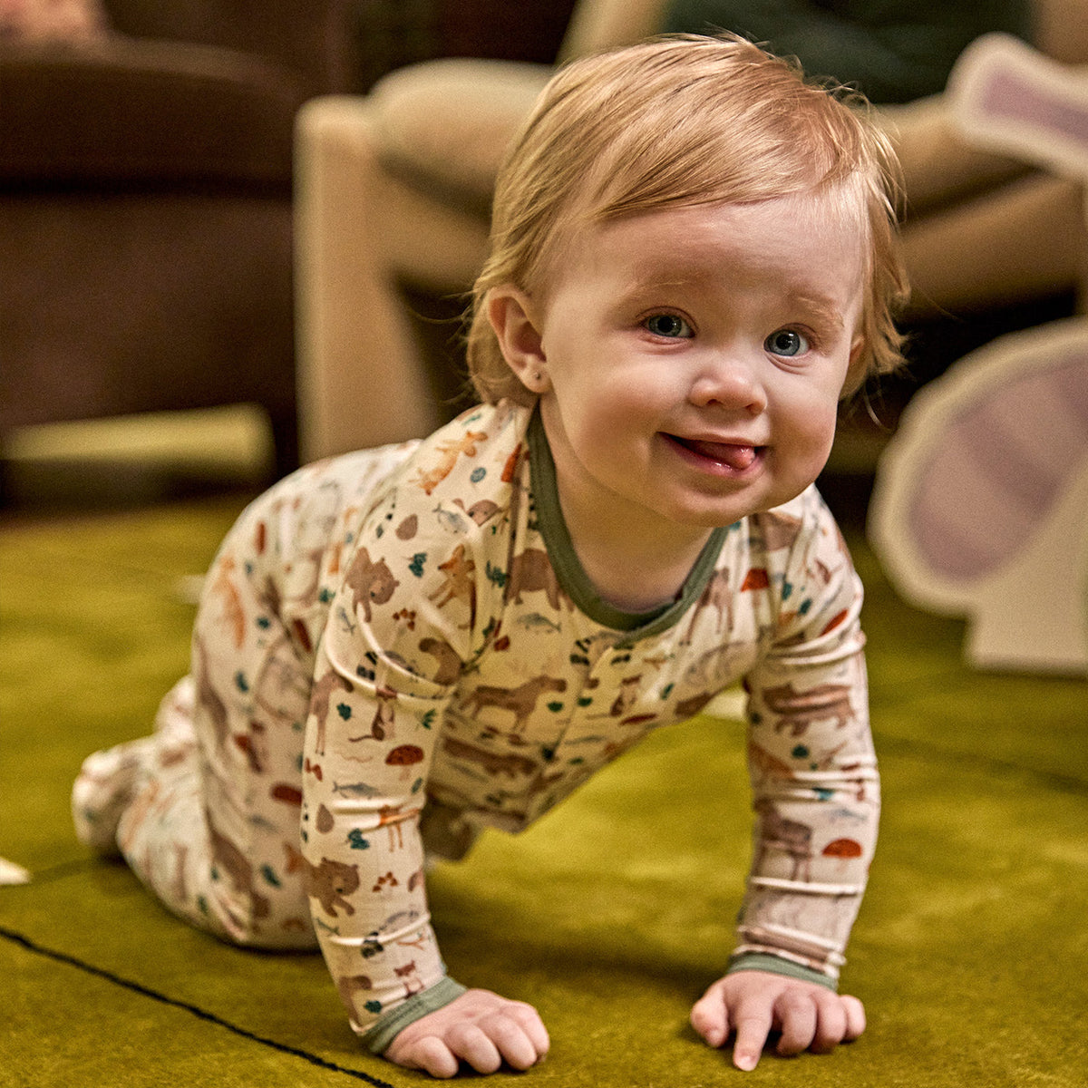 A smiling baby crawling on a green carpet, wearing a long-sleeve footed pajama top with a safari animal print on a cream background.