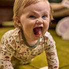 A close-up of a baby crawling on a green carpet, wearing a cream-colored long-sleeve pajama top with a colorful animal print and a green collar.