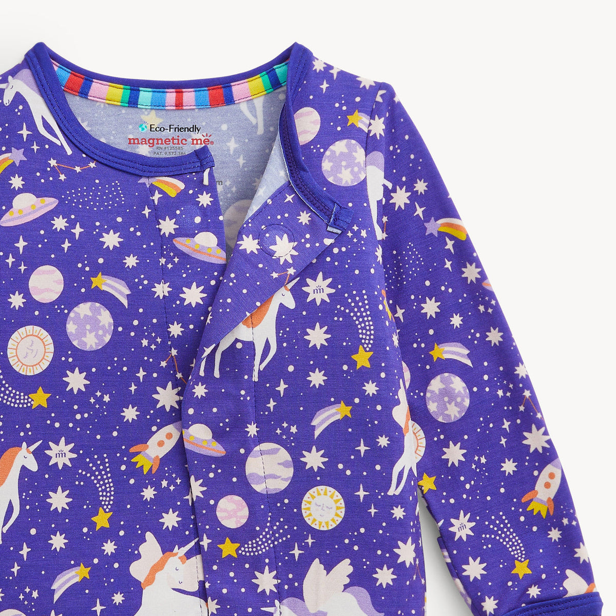 A close-up of a baby long-sleeve sleep sack or pajama top in purple, featuring a white and pastel multicolor space-themed print with unicorns, rockets, planets, and stars. The inner collar has a colorful striped trim, and the inside neck label reads 'Eco-Friendly magnetic ink baby'.