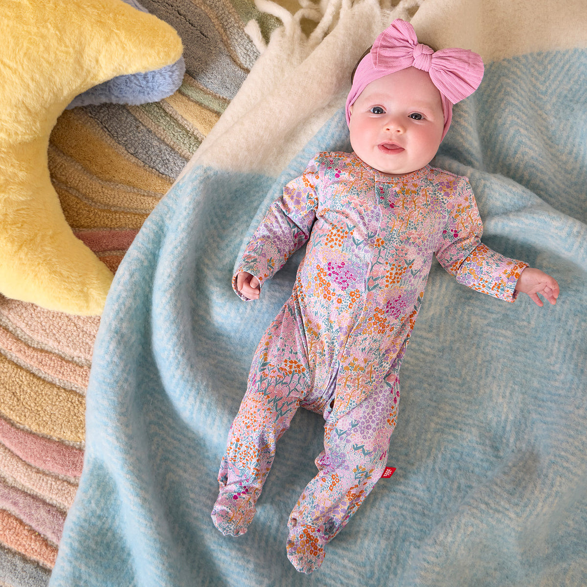 A baby lying on a light blue blanket, wearing a pink footed sleeper with a colorful floral print and a matching pink bow headband.