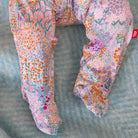 A close-up of a pair of patterned pants laid flat on a textured blue-gray surface. The fabric has an all-over floral and botanical print in pink, purple, orange, teal, and white, with a small red brand tag visible on the upper right leg.