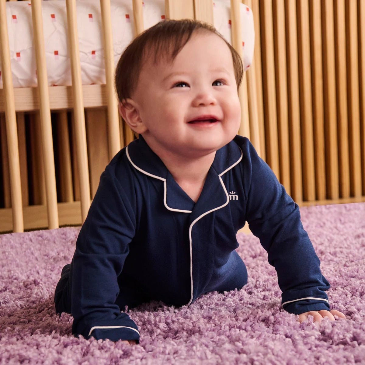 A baby crawling on a purple shag rug in front of a wooden crib, wearing a navy blue long-sleeve pajama top with white piping and a small embroidered monogram logo.