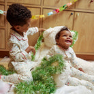 Two young children wearing matching holiday pajamas sit together on a white plush rug in front of wooden cabinets, surrounded by green tinsel garland and a white teddy bear.