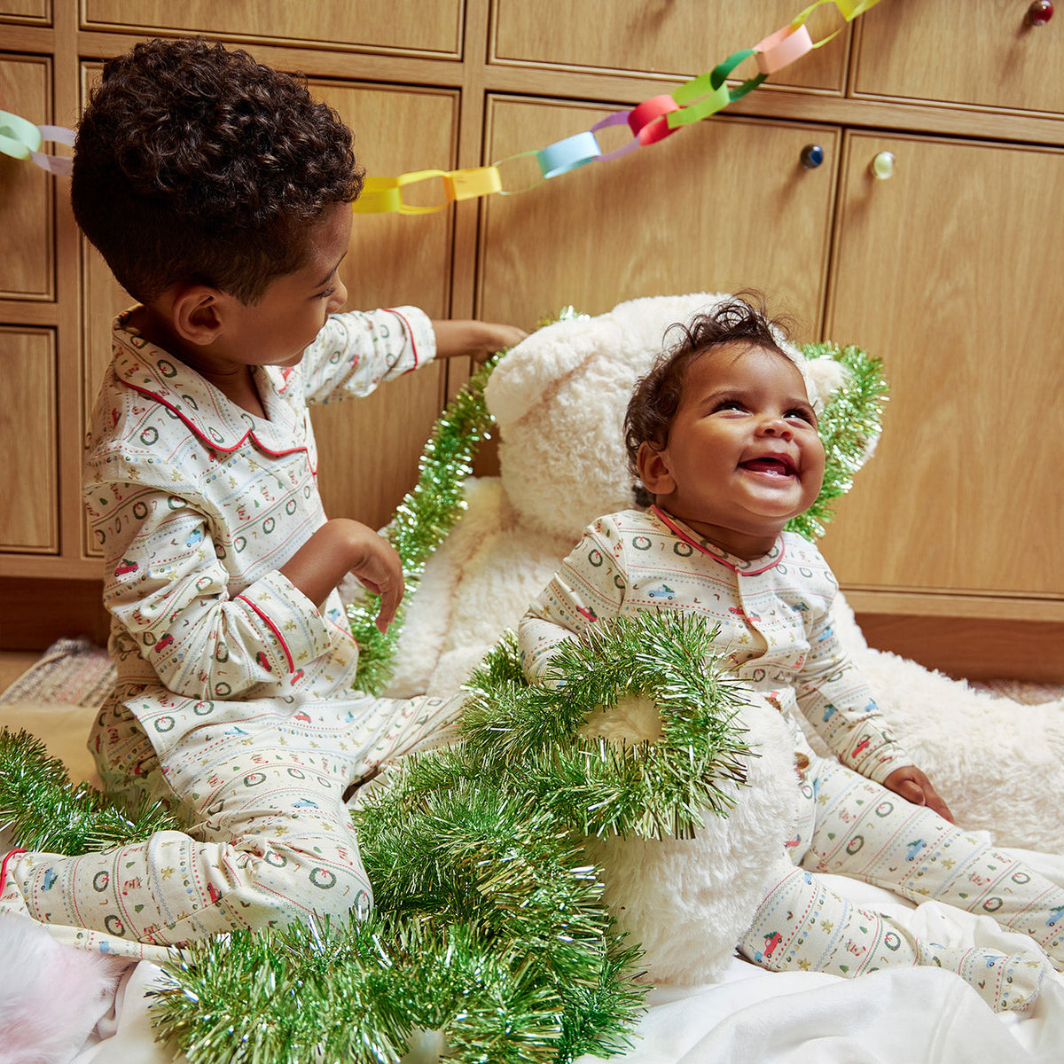 Two young children wearing matching holiday pajamas sit together on a white plush rug in front of wooden cabinets, surrounded by green tinsel garland and a white teddy bear.