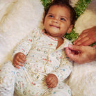 A baby wearing a cream-colored long-sleeve pajama set with a colorful holiday pattern, lying on a white fluffy blanket with a hand adjusting the collar. The baby is also wearing a green tinsel headband.