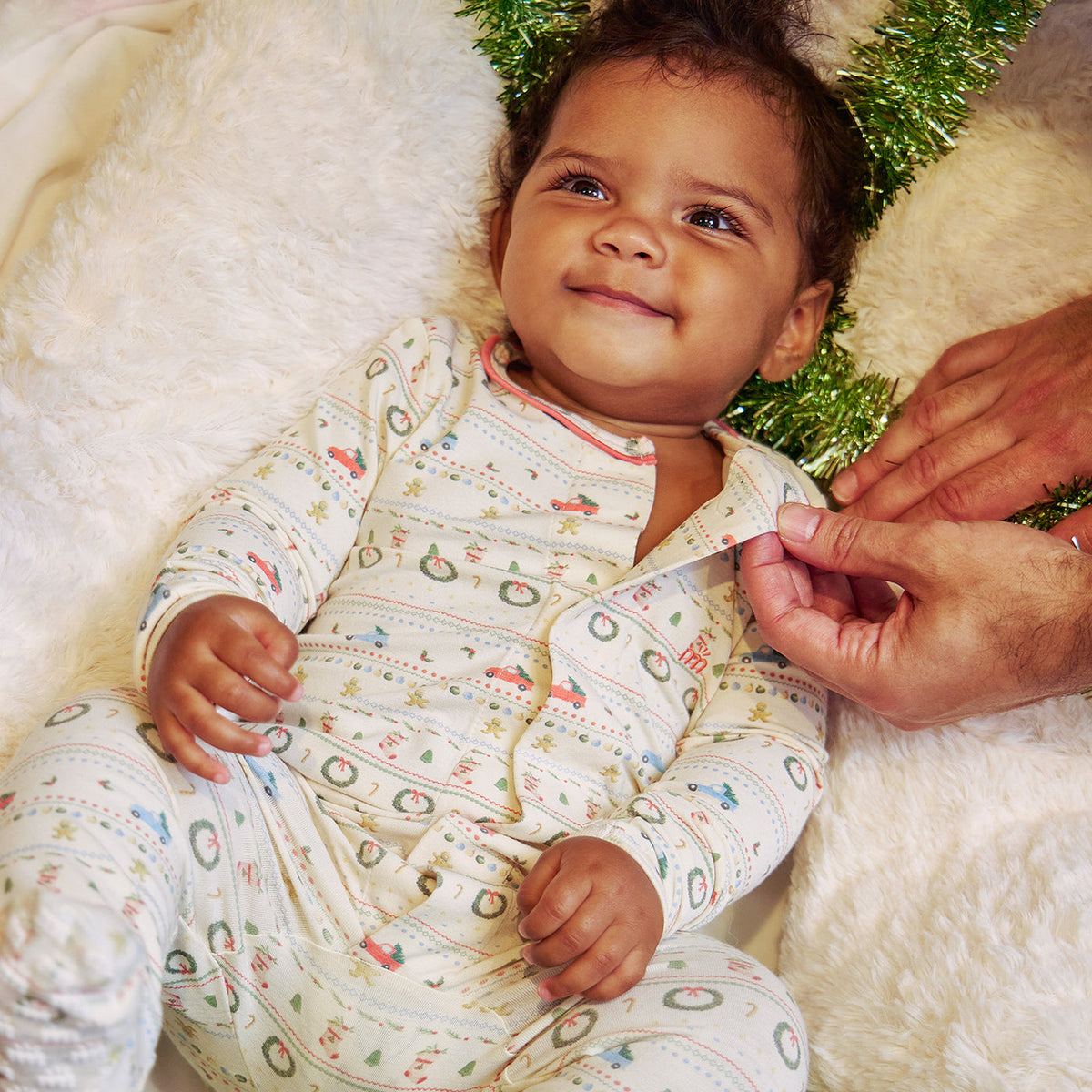 A baby wearing a cream-colored long-sleeve pajama set with a colorful holiday pattern, lying on a white fluffy blanket with a hand adjusting the collar. The baby is also wearing a green tinsel headband.