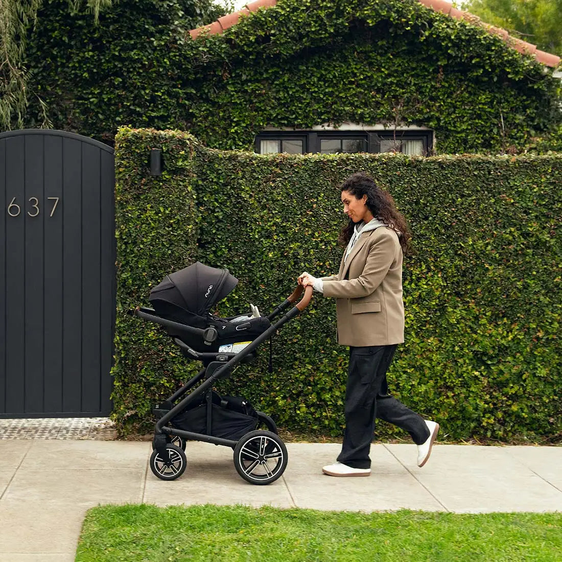 A woman with long dark hair pushing a black MIXX Next + Pipa Aire RX Travel System by Nuna stroller along a sidewalk past a dark gate and a vine-covered wall.