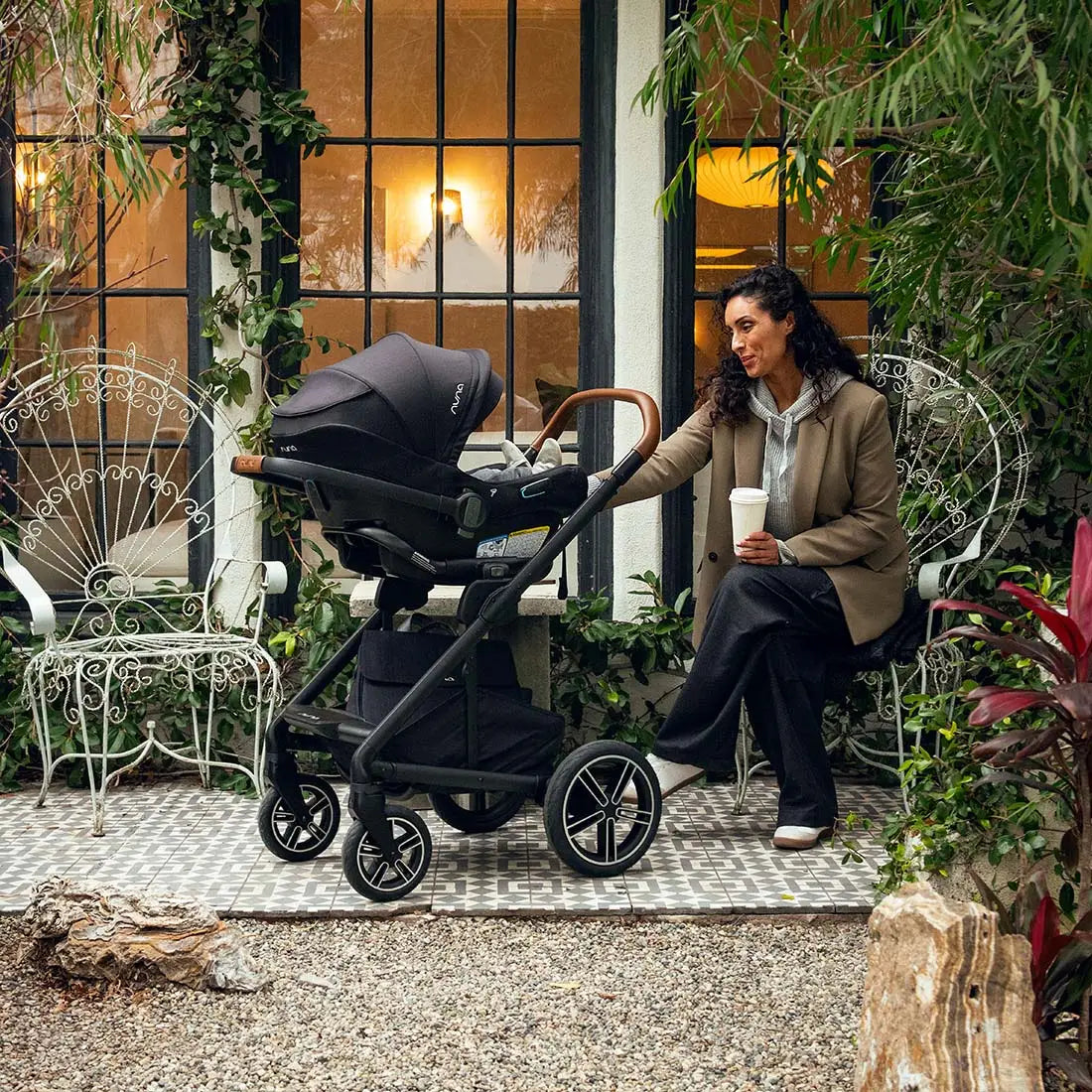 A woman sitting in a courtyard next to a black MIXX Next + Pipa Aire RX Travel System by Nuna stroller holding a cup.