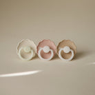Three baby pacifiers in white, pink, and beige on a beige background