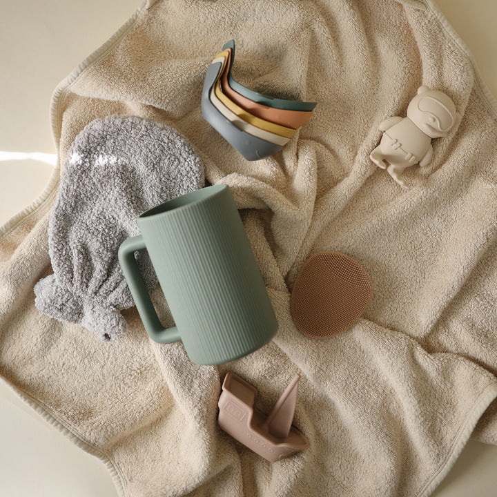 A set of stacked toy whales, a green ceramic mug, a textured washcloth, a round silicone sponge, and a small toy figure arranged on top of a soft, textured cream-colored blanket.