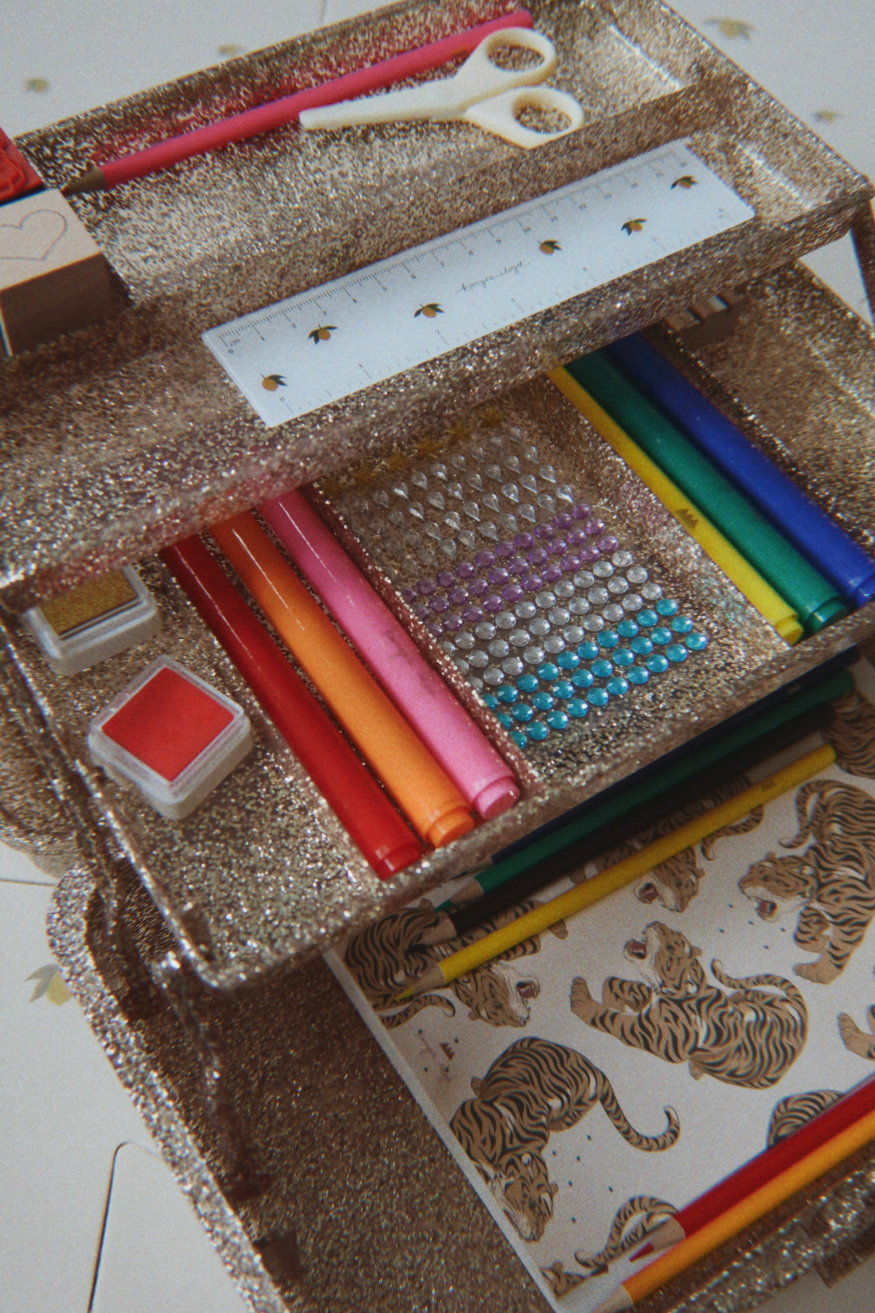A glittery gold tiered storage organizer filled with colorful art supplies, including markers, colored pencils, a ruler, scissors, rhinestones, and a tiger-themed drawing sheet.