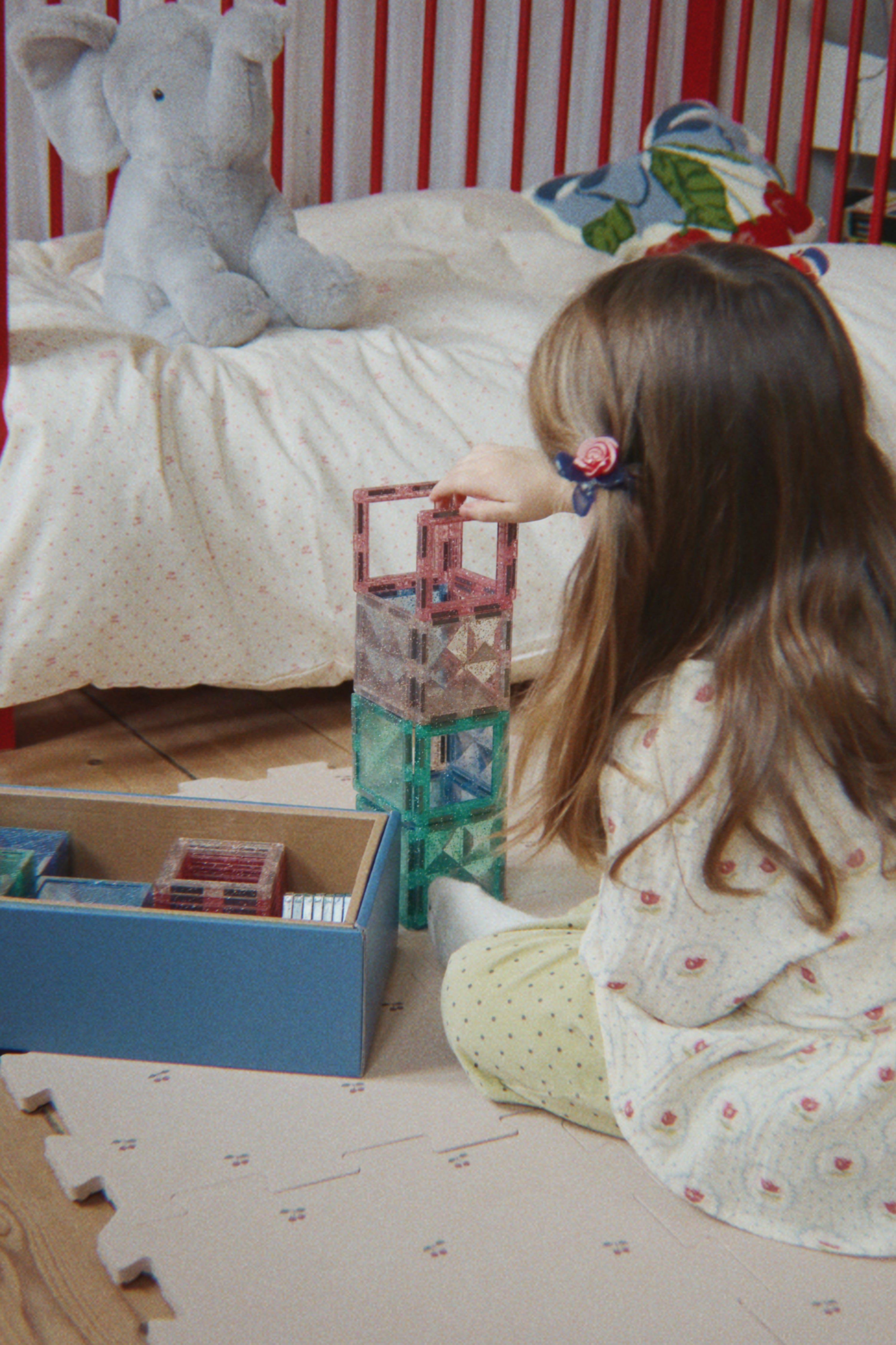 A young girl with long brown hair sits on a play mat in a nursery, building a tower with translucent plastic blocks. A blue storage box with more blocks is on the floor, and a white crib with red stripes and a gray stuffed elephant is in the background.