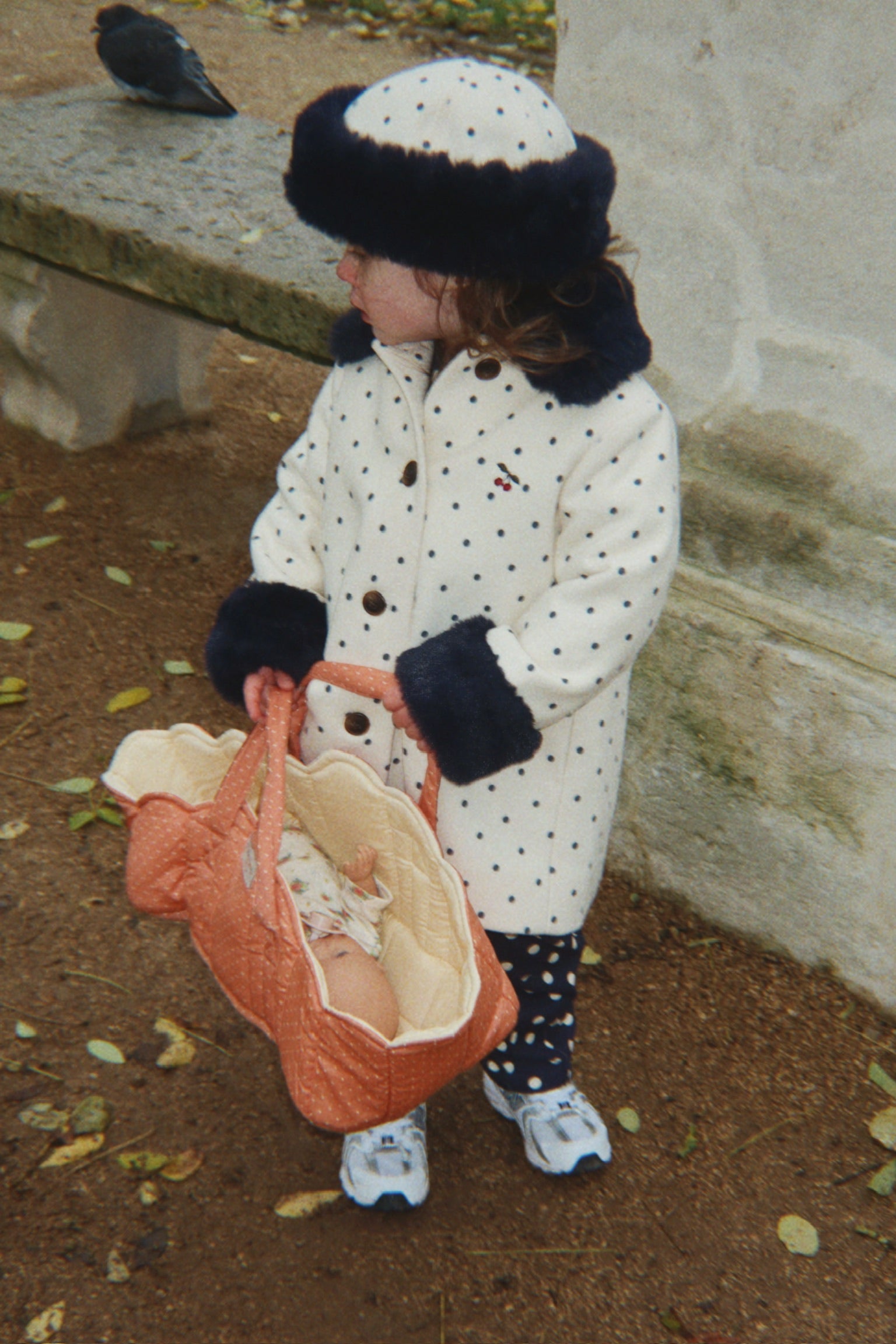 A toddler standing outdoors in a cream winter coat with black polka dots and dark fur trim, holding a baby in an orange bassinet. The child also wears a matching cream hat with dark trim and white sneakers.