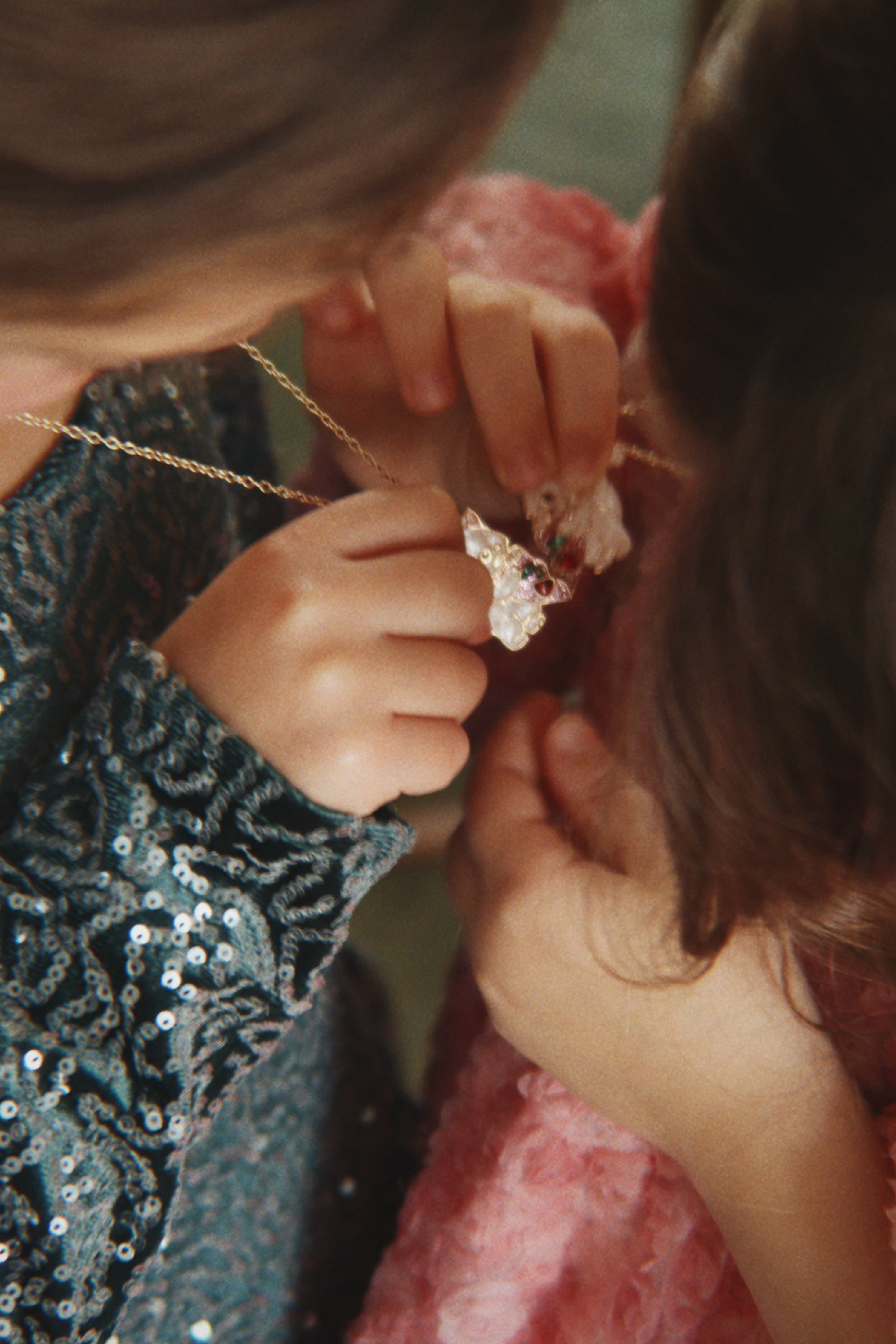 A close-up lifestyle shot of a child wearing a sparkling teal sequined top while another person adjusts a delicate gold necklace with a small white flower pendant.