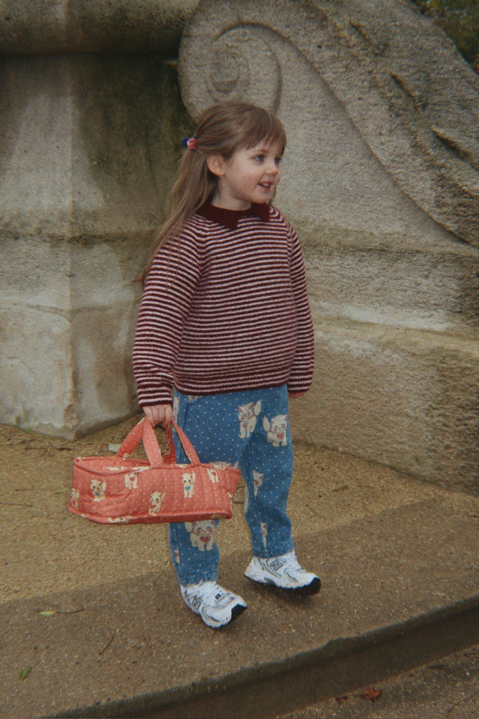 Young girl in a striped sweater and floral pants holding a red bag against a stone wall.