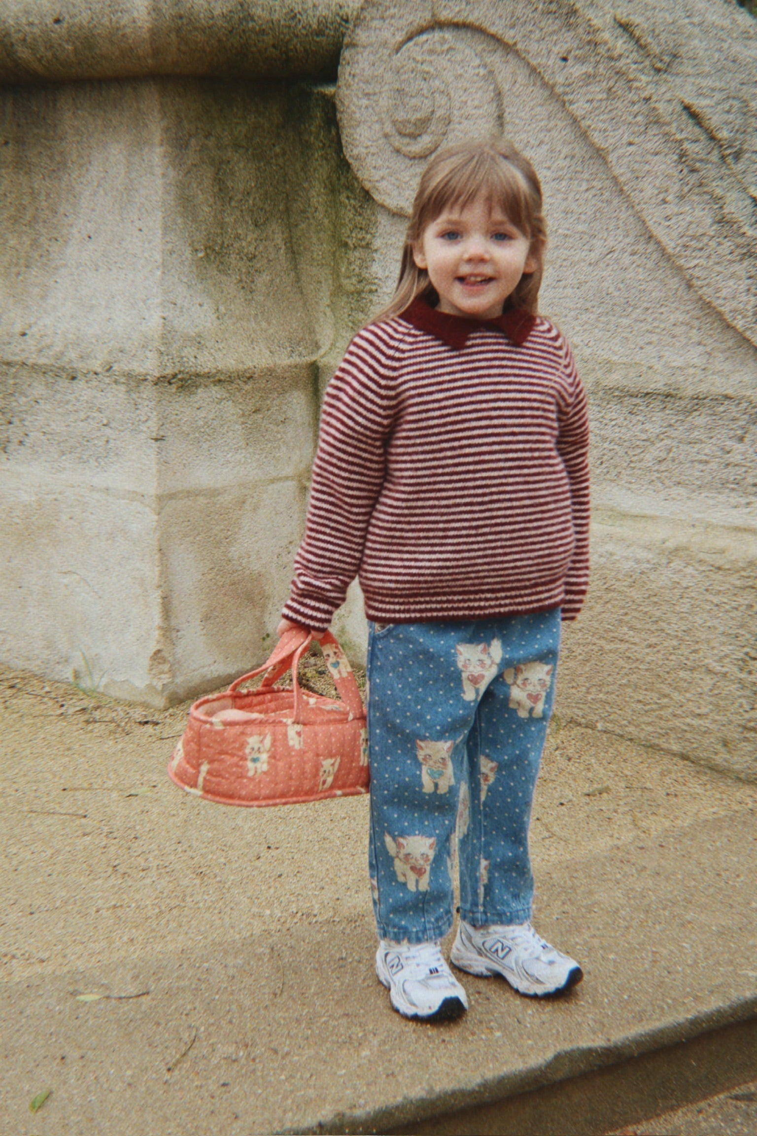 A young girl with light brown hair stands outdoors in front of a stone wall, wearing a red and white striped long-sleeve sweater, blue jeans with a teddy bear print, and white sneakers. She is holding a coral pink patterned handbag.
