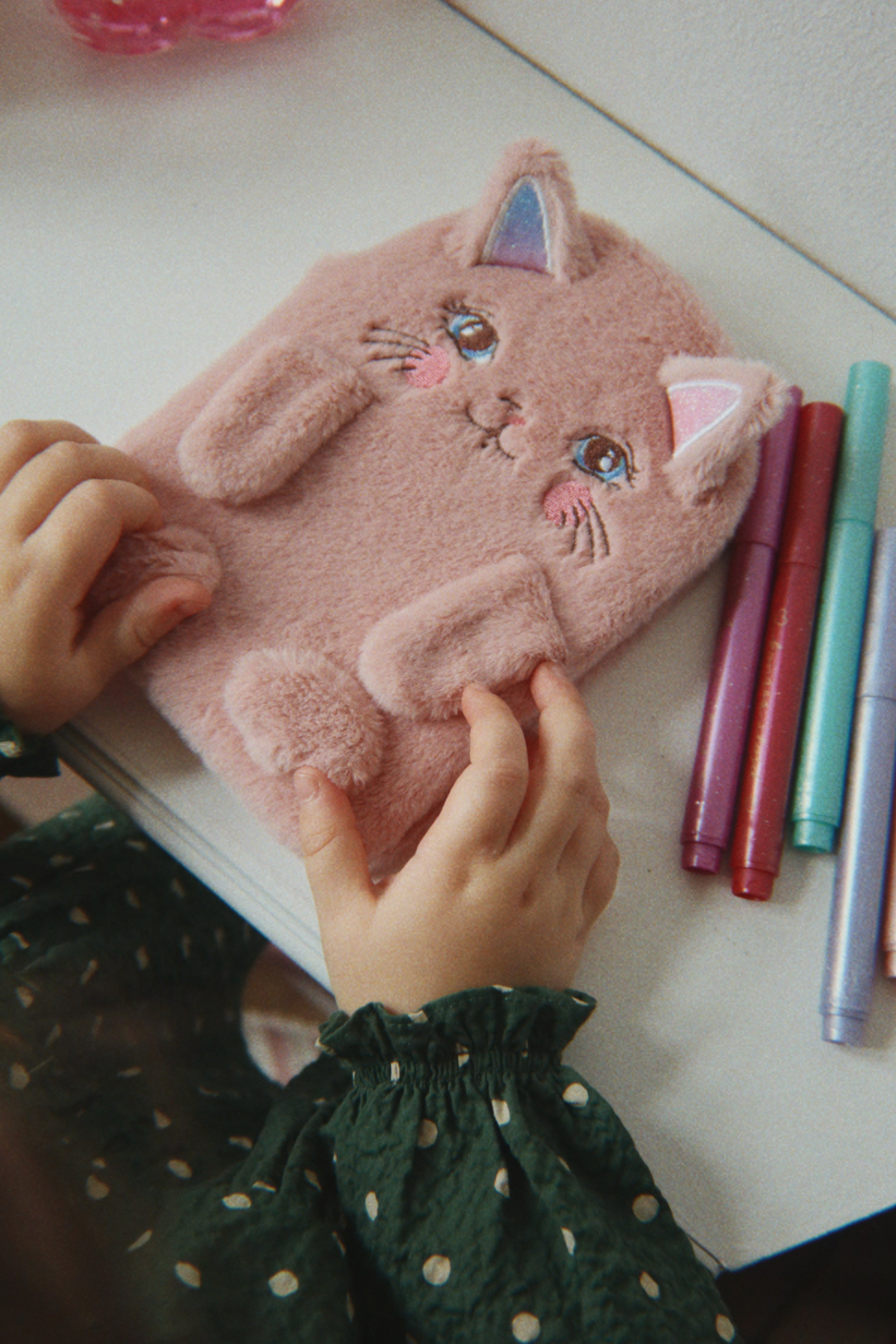 A child's hands hold a pink fuzzy cat-shaped notebook or journal on a white desk, with colorful markers nearby.