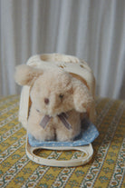 Small plush toy with a bow on a decorative stand against a white curtain background