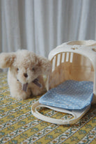 A small beige plush dog toy sits beside a cream-colored pet carrier with a blue polka-dot cushion, placed on a yellow patterned surface against a soft gray curtain background.