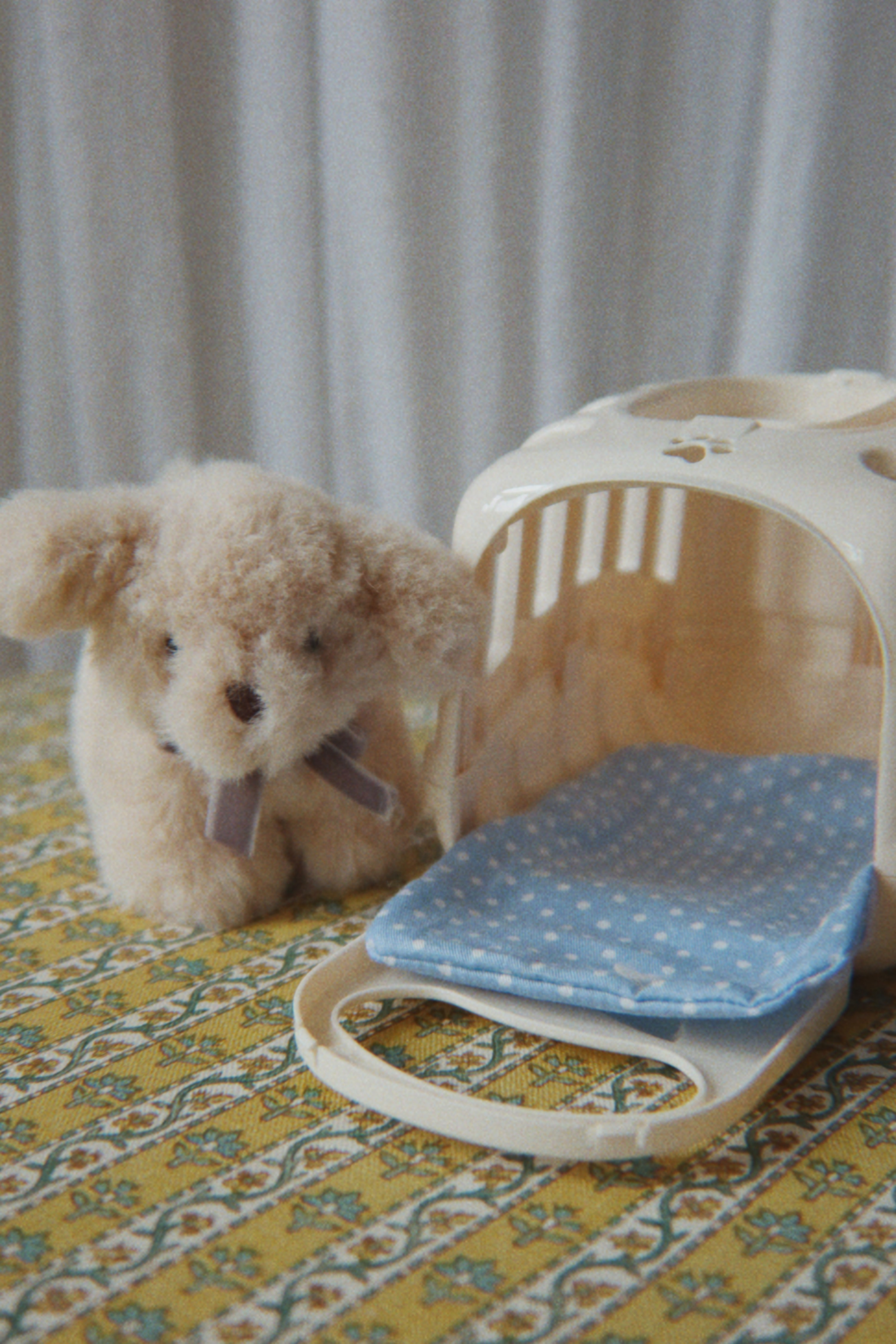 A small beige plush dog toy sits beside a cream pet carrier with a blue polka-dot cushion, placed on a yellow floral-patterned surface against a soft gray curtain background.