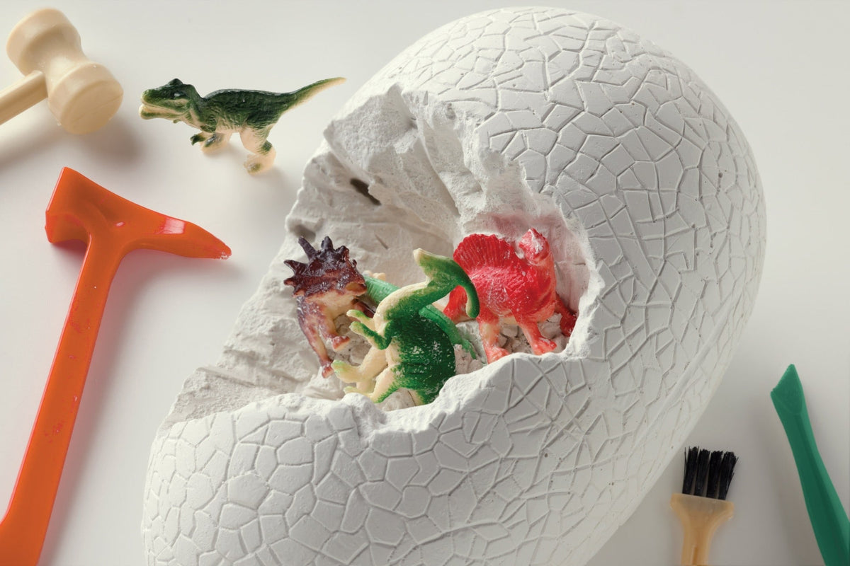 A large, cracked white dinosaur egg revealing small toy dinosaurs nestled inside, surrounded by an orange mallet, a small green brush, and other excavation tools on a white background.