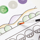 A close-up of a hand holding a fine paintbrush and coloring a printed illustration. The artwork shows outlined shapes and watercolor circles in green, lavender, peach, gray, and purple on a white background.