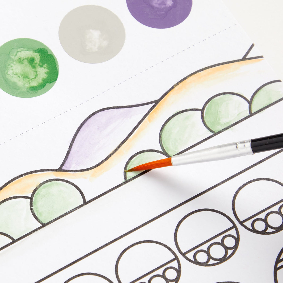 A close-up of a hand holding a fine paintbrush and coloring a printed illustration. The artwork shows outlined shapes and watercolor circles in green, lavender, peach, gray, and purple on a white background.
