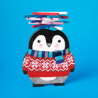 Wooden game piece for the Penguin Stack-up Wooden Balancing Game, shaped like an illustrated penguin with rosy cheeks, wearing a festive red patterned fair isle sweater and a blue-and-green striped scarf, with a balanced stack of colorful wooden sticks on its head against a bright blue background.
