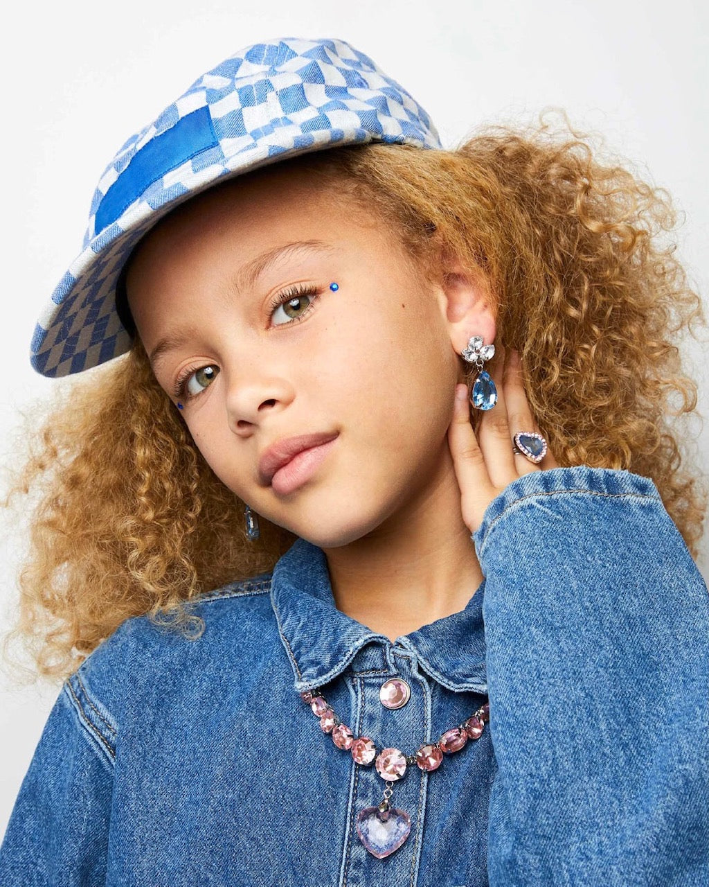 Young girl wearing a blue denim jacket, checkered hat, and jewelry against a white background