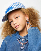 Young girl wearing a blue denim jacket, checkered hat, and jewelry against a white background