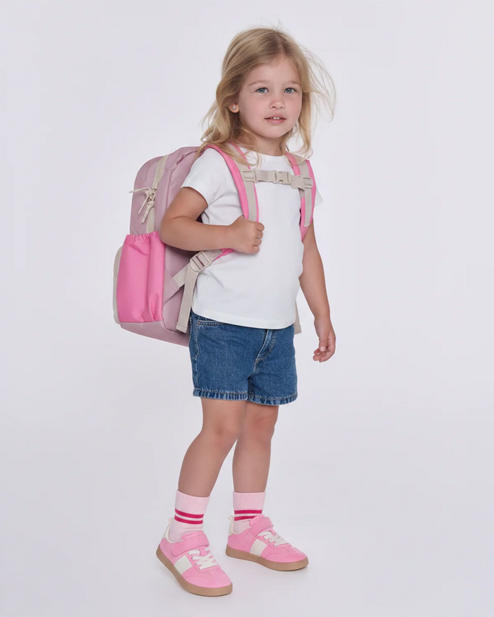 A young girl with blonde hair stands against a light gray studio background wearing a pink backpack, a white t-shirt, denim shorts, pink socks, and pink shoes.