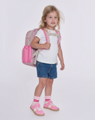 A young girl with blonde hair stands against a light gray studio background wearing a pink backpack, a white t-shirt, denim shorts, pink socks, and pink shoes.