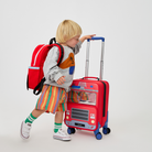 A toddler standing between a red State Bags Mini Logan firetruck-style backpack and a matching red rolling carry-on suitcase, wearing a gray sweatshirt with an orange graphic and striped socks.