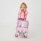A child standing behind a pink State Bags Mini Logan house-style rolling carry-on suitcase and matching backpack, wearing a pink jacket with red trim.