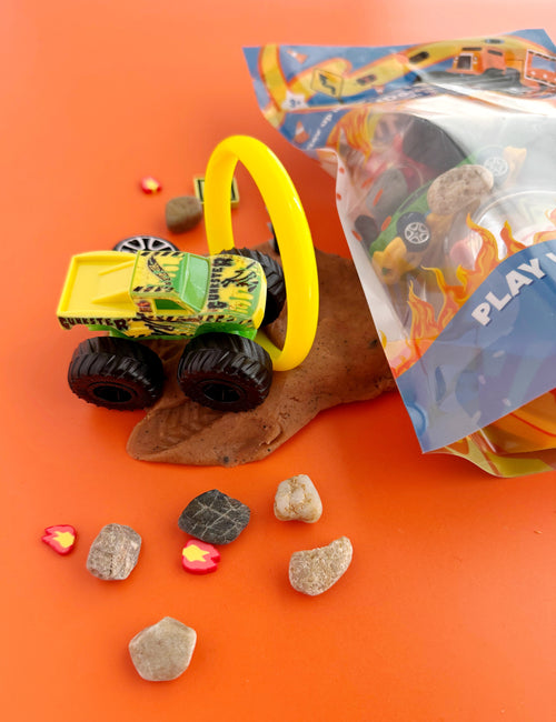 Toy truck with a yellow handle on an orange surface with rocks and a bag labeled 'Play' in the background.