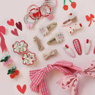 A styled flat-lay of cute pink and red hair accessories on a light pink background, including fabric hair bows, hair clips, and small decorative hair charms.