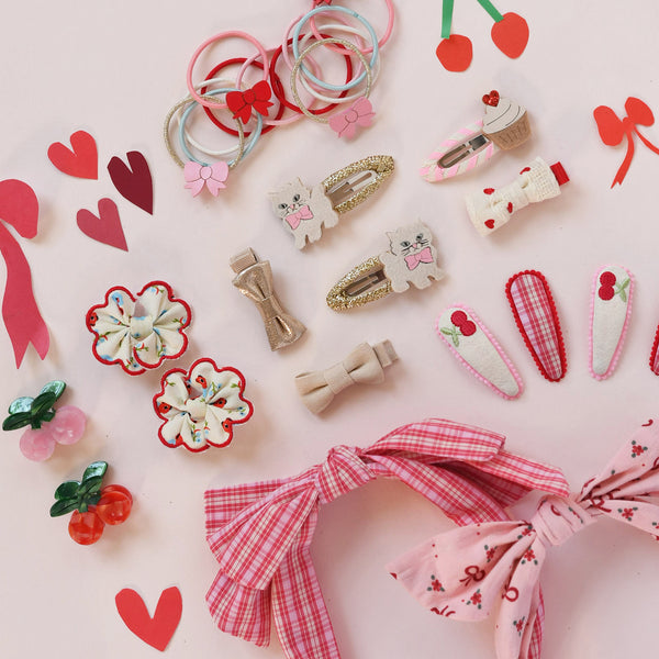 A styled flat-lay of cute pink and red hair accessories on a light pink background, including fabric hair bows, hair clips, and small decorative hair charms.