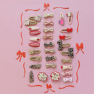 A flat-lay collection of 24 cute hair clips arranged on a solid pink background, featuring various patterns like leopard print, hearts, bows, and cherries in pastel and red tones.