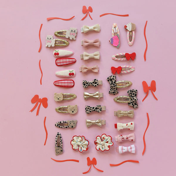 A flat-lay collection of 24 cute hair clips arranged on a solid pink background, featuring various patterns like leopard print, hearts, bows, and cherries in pastel and red tones.
